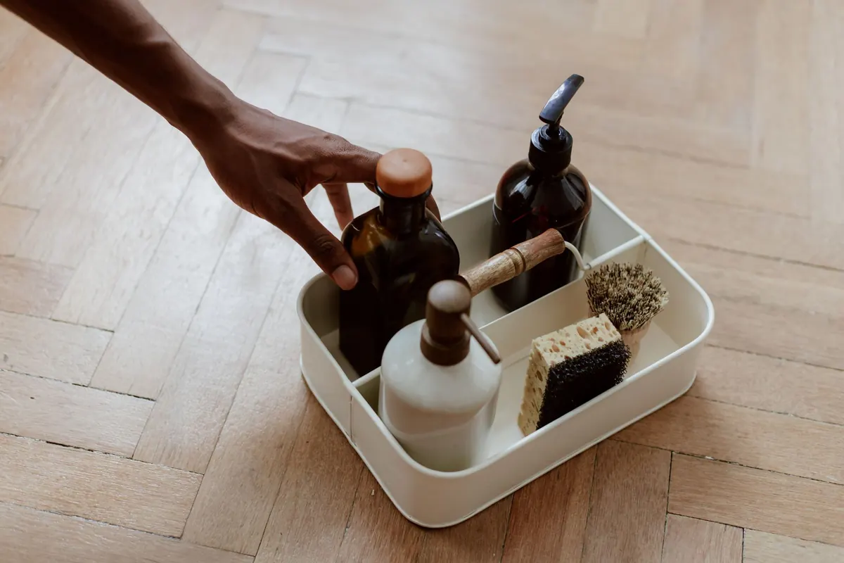 A hand reaching into a cleaning caddy of bottles, brushes and a sponge on a wooden floor
